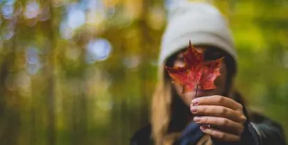 person holding red maple leaf in front of face with blurred trees in background